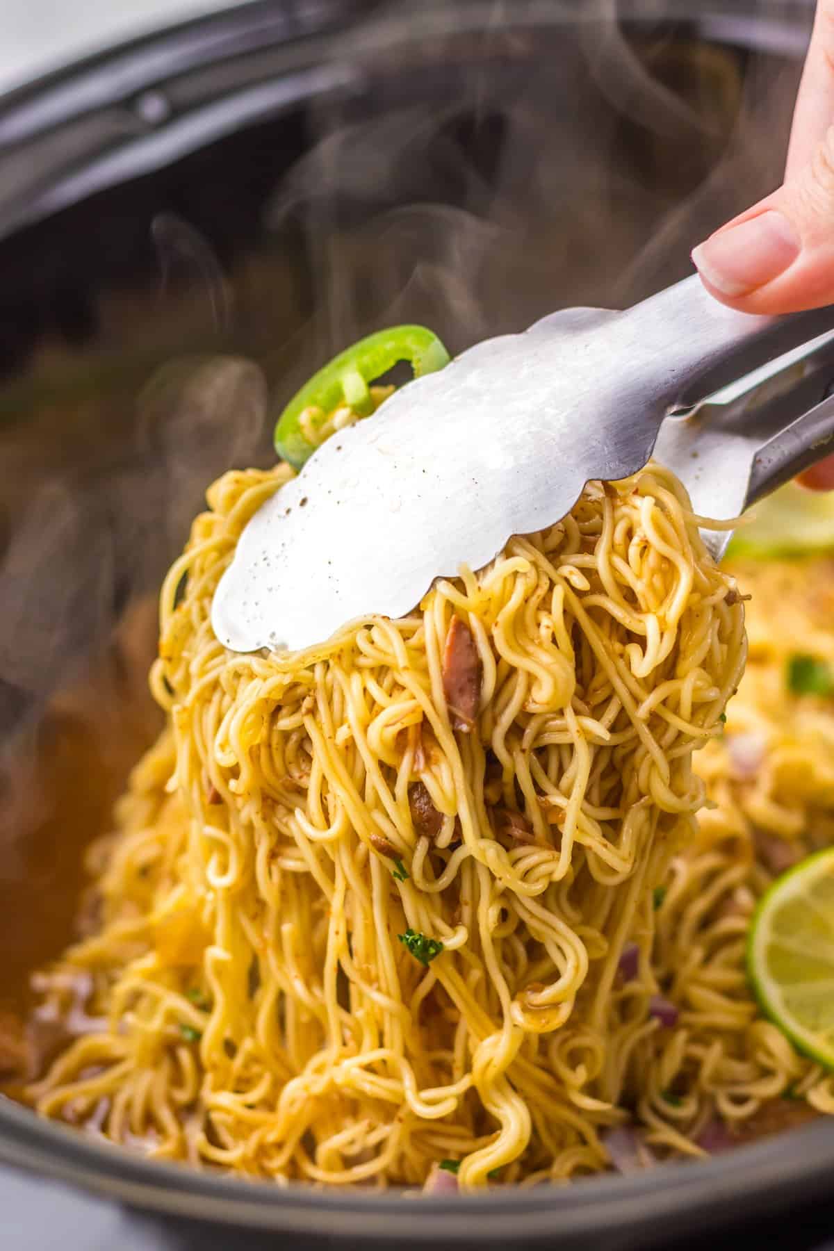 Tender birria beef slow cooks all day before getting tossed with ramen noodles and toppings in this Birria Ramen recipe. Tongs grabbing birria ramen from the slow cooker