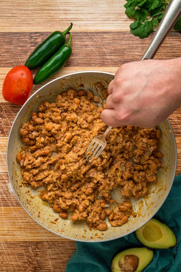 Crunchy tostada shells are topped with homemade refried beans, veggies, avocado, and cheese in this easy Tostada Recipe. A hand using a fork to mash beans in a pan