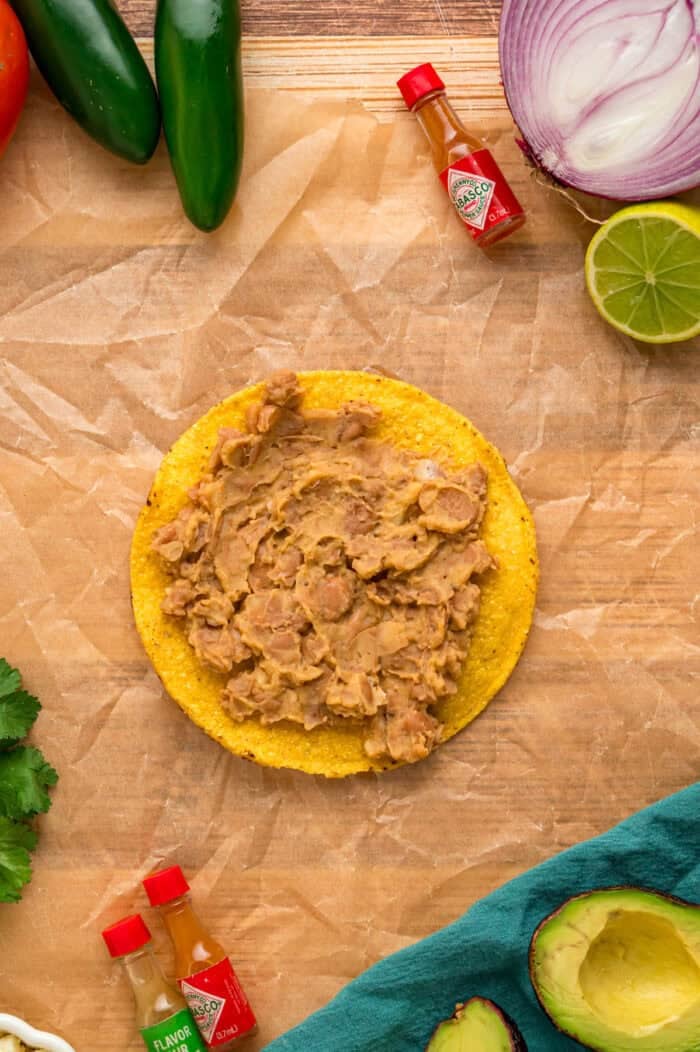 Crunchy tostada shells are topped with homemade refried beans, veggies, avocado, and cheese in this easy Tostada Recipe. Refried beans spread on a tostada shell